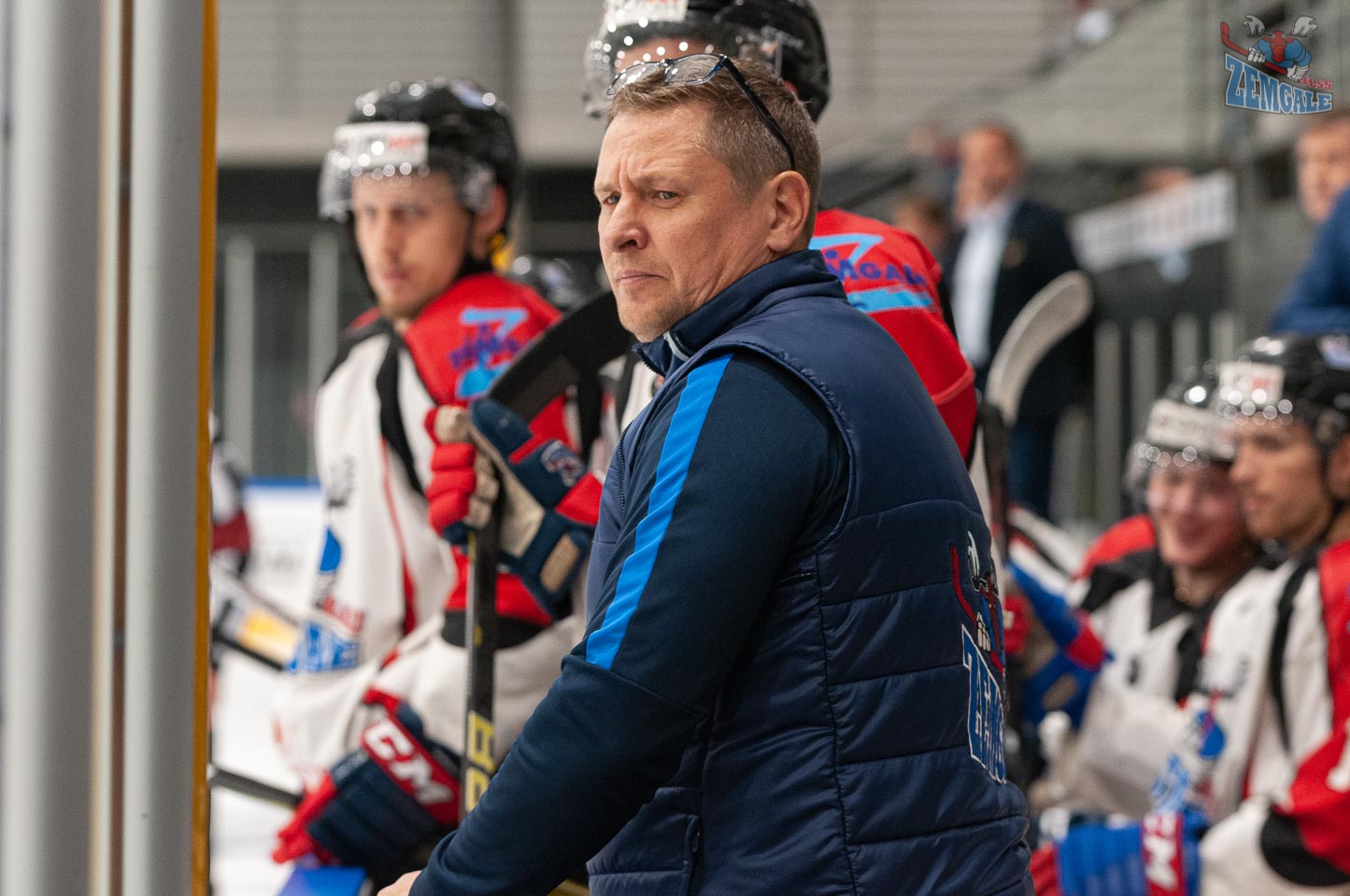 Vīrietis saviebi seju baidoties no ripas lidojuma, kamēr viņam aizmugurē stāv hokejisti